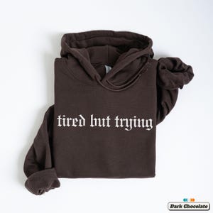 May include: A dark brown hoodie with the text "tired but trying" in white gothic font.