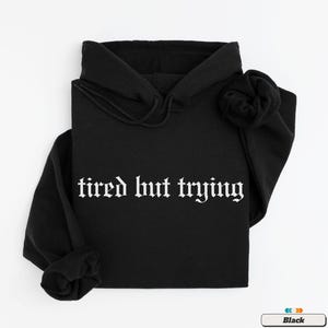 May include: Black hooded sweatshirt with the text "tired but trying" in white gothic font.