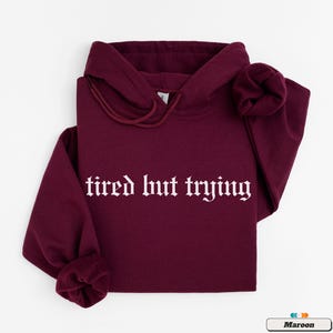 May include: A maroon hoodie with the text "tired but trying" in white gothic font.