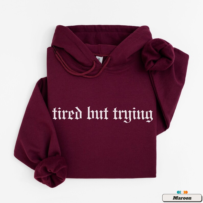 May include: A maroon hoodie with the text "tired but trying" in white gothic font.