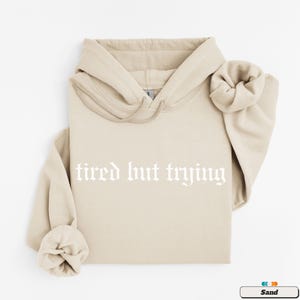May include: A sand-colored hoodie with a white drawstring and the text "tired but trying" in a gothic font.