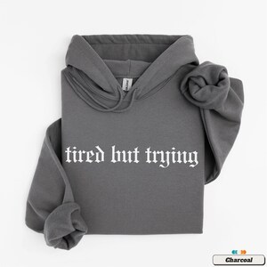 May include: A charcoal gray hooded sweatshirt with the text "tired but trying" in white gothic font.