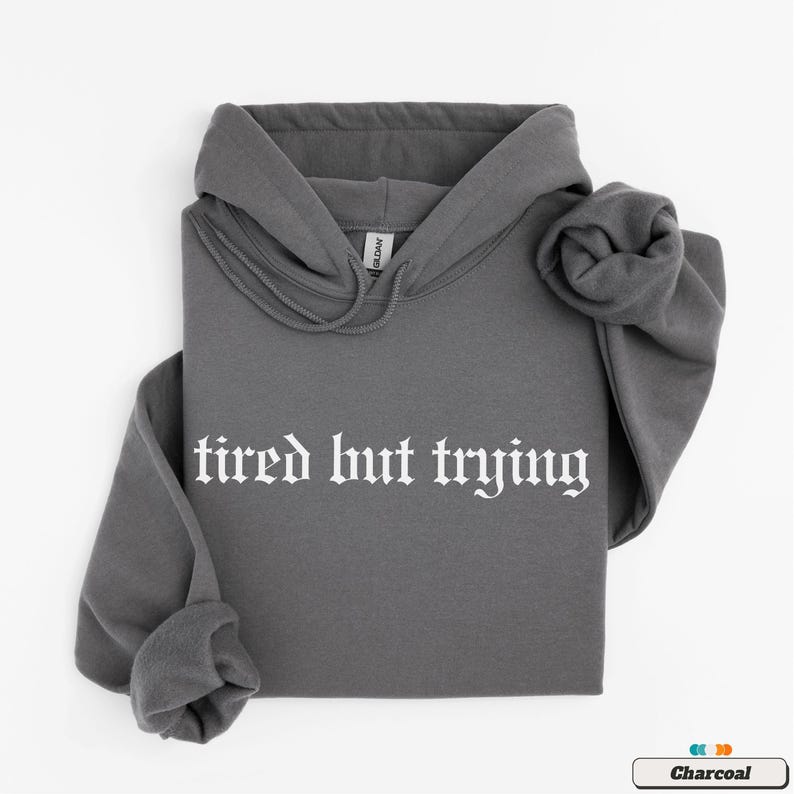 May include: A charcoal gray hooded sweatshirt with the text "tired but trying" in white gothic font.