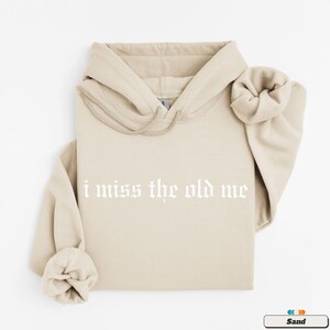 Puede incluir: Una sudadera con capucha de color arena con un cord&oacute;n blanco y el texto "i miss the old me" impreso en blanco en la parte delantera.