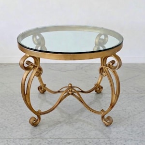 May include: A round side table with a clear glass top and a gold-colored metal base. The base features ornate scrollwork and curved legs. The table is approximately 24 inches in diameter and 20 inches tall.