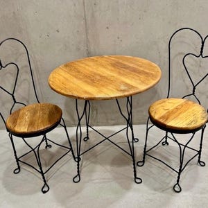 May include: A small bistro set with a round wooden tabletop and two chairs. The chairs and table base are black wrought iron. The chair backs feature a heart design. The seats are made of wood.