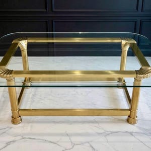 May include: A rectangular coffee table with a clear glass top and a gold-colored metal frame. The frame features decorative shell-like accents at the corners and along the legs. The table is supported by four cylindrical legs.