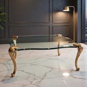 May include: A clear glass coffee table with ornate gold-colored legs. The table has a rectangular shape with curved edges. The legs feature a detailed, scrolled design, adding a touch of elegance to the furniture.
