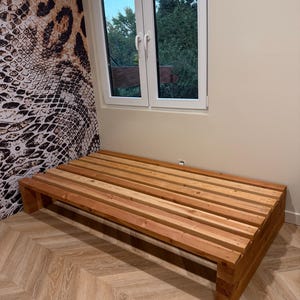 May include: A low-profile platform bed frame constructed from natural light brown wooden slats. The rectangular frame features a slatted surface design supported by sturdy block-style legs at each corner, providing a simple and functional base.