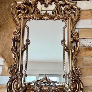 May include: Ornate gold-colored mirror with a rectangular reflective surface. The frame features elaborate scrollwork and floral details, creating a vintage aesthetic. The mirror is designed for wall mounting and adds a touch of elegance to any room.
