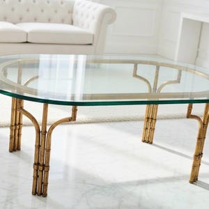 May include: Oval glass-top coffee table with a gold-colored bamboo-style base. The table has a clear glass top and a decorative gold trim. The legs are made to look like bamboo stalks. The table is in a bright, well-lit room.