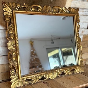 May include: A rectangular mirror with an ornate, gold-colored frame. The frame features detailed scrollwork and a decorative design. The mirror reflects a room with a Christmas tree and a window. The mirror is placed on a wooden surface.