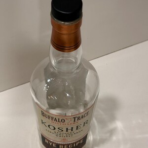 K&ouml;nnte beinhalten: Klare Glasflasche Buffalo Trace Kosher Rye Recipe Whiskey. Die Flasche hat einen schwarzen Verschluss und einen kupferfarbenen Hals. Das Etikett hat Text in Wei&szlig; und Braun.