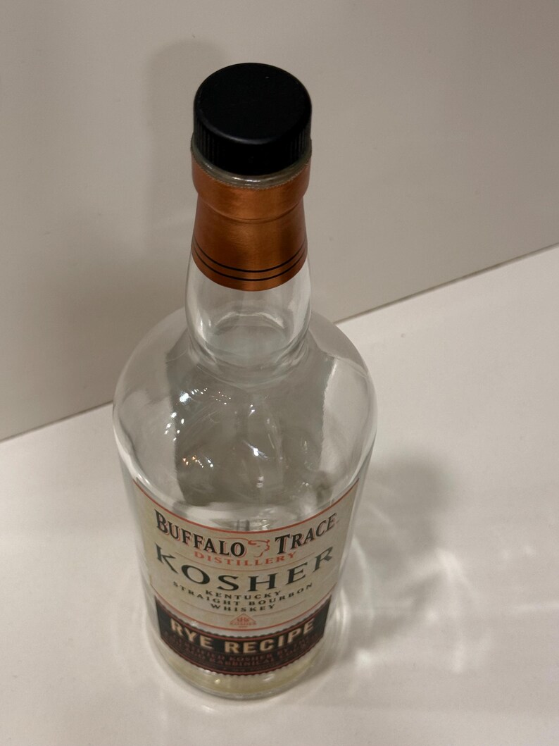 K&ouml;nnte beinhalten: Klare Glasflasche Buffalo Trace Kosher Rye Recipe Whiskey. Die Flasche hat einen schwarzen Verschluss und einen kupferfarbenen Hals. Das Etikett hat Text in Wei&szlig; und Braun.