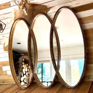 May include: Three oval mirrors with ornate gold-colored frames. The mirrors are arranged in a row, reflecting the surrounding room. The frames have a detailed, textured design. The mirrors are placed on a wooden surface, with a rustic wooden wall in the background.