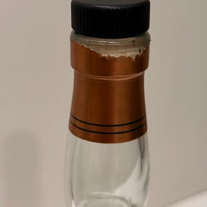 K&ouml;nnte beinhalten: Eine klare Glasflasche mit schwarzem Verschluss und kupferfarbener Folienversiegelung. Die Folienversiegelung hat zwei schwarze Streifen und ist teilweise zerrissen. Die Flasche ist leer.