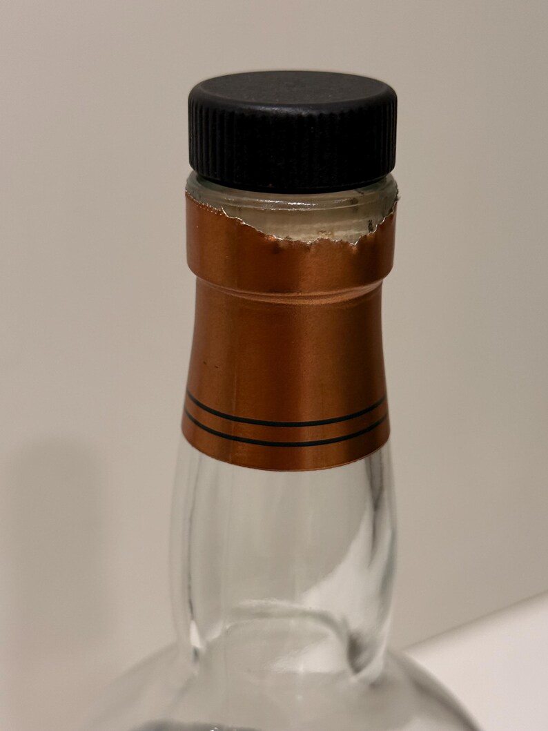 K&ouml;nnte beinhalten: Eine klare Glasflasche mit schwarzem Verschluss und kupferfarbener Folienversiegelung. Die Folienversiegelung hat zwei schwarze Streifen und ist teilweise zerrissen. Die Flasche ist leer.