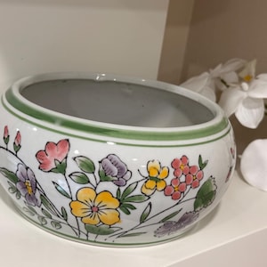 May include: A round white ceramic bowl featuring a hand-painted floral pattern with yellow, pink, purple, and green flowers. The rim is accented with a thin green band, and the interior of the bowl is a smooth, plain white finish.