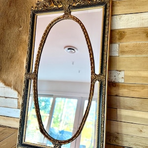 May include: Ornate rectangular mirror with a gold and black frame. The mirror features an oval gold accent in the center and decorative carvings at the top and corners. The mirror is displayed on a wooden surface against a wood-paneled wall.