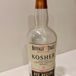 May include: Clear glass bottle of Buffalo Trace Kosher Kentucky Straight Bourbon Whiskey. The bottle has a black cap and a copper-colored neck. The label reads "Rye Recipe" and "Certified Kosher by the Chicago Rabbinical Council."