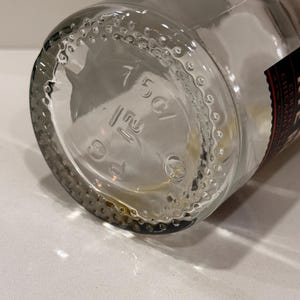 K&ouml;nnte beinhalten: Klare Glasflasche mit rundem Boden. Der Boden der Flasche hat gepr&auml;gte Markierungen, darunter "150C/". Der Boden ist von kleinen, erhabenen Punkten umgeben. Ein Teil eines Etiketts ist sichtbar.