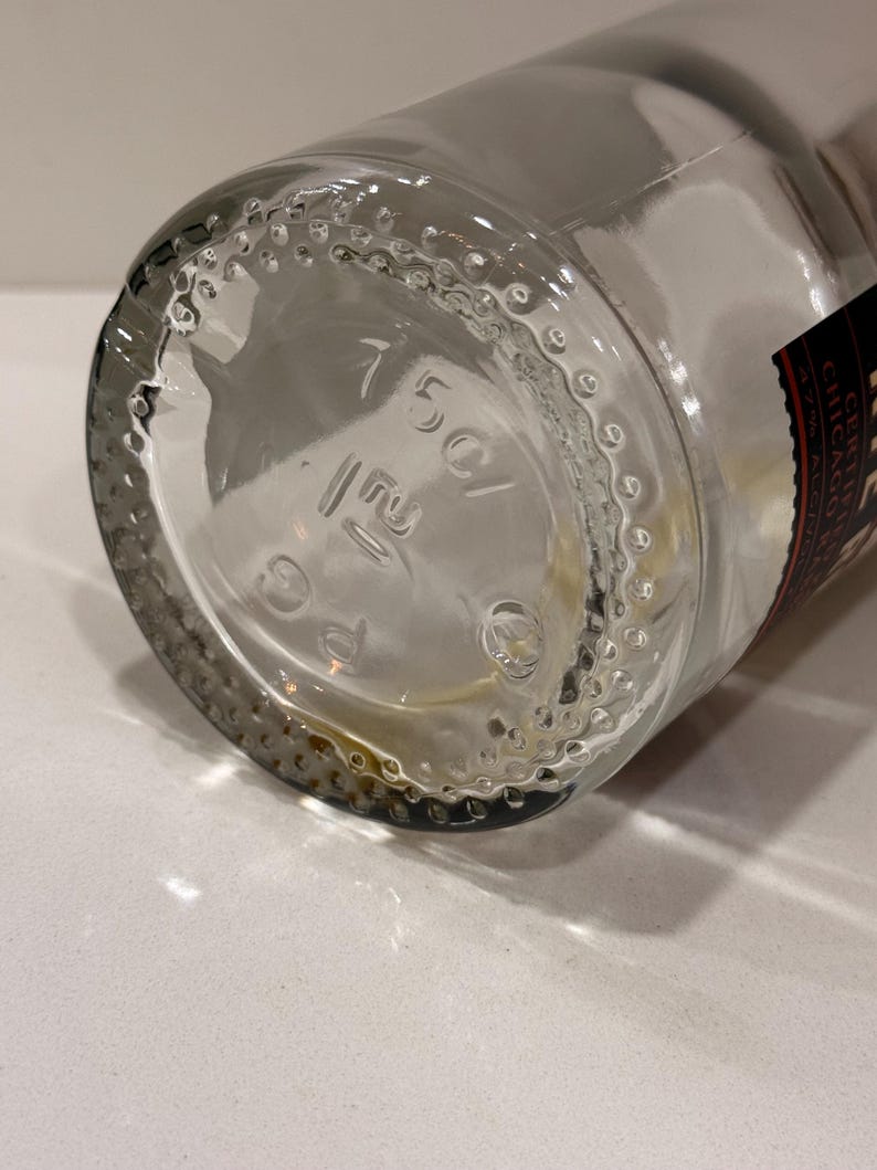 K&ouml;nnte beinhalten: Klare Glasflasche mit rundem Boden. Der Boden der Flasche hat gepr&auml;gte Markierungen, darunter "150C/". Der Boden ist von kleinen, erhabenen Punkten umgeben. Ein Teil eines Etiketts ist sichtbar.