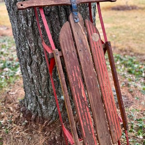 May include: An antique wooden sled with red metal runners and accents leans against a tree. The sled's wooden slats are painted with red stripes, showing signs of wear. A classic winter recreational item.