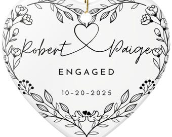 Może przedstawiać: Biała ceramiczna ozdoba w kształcie serca ze złotym sznurkiem do zawieszenia. Ozdoba zawiera imiona "Robert" i "Paige", słowo "ENGAGED" oraz datę "10-20-2025". Wzory kwiatowe i liściaste otaczają tekst.