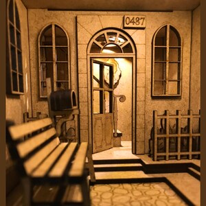 Peut inclure: Mod&egrave;le en bois d'une maison avec une porte, deux fen&ecirc;tres et une bo&icirc;te aux lettres. La maison porte le num&eacute;ro "0487" au-dessus de la porte. Il y a un banc en bois devant la maison.