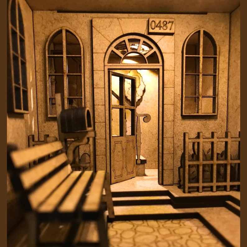 Peut inclure: Mod&egrave;le en bois d'une maison avec une porte, deux fen&ecirc;tres et une bo&icirc;te aux lettres. La maison porte le num&eacute;ro "0487" au-dessus de la porte. Il y a un banc en bois devant la maison.