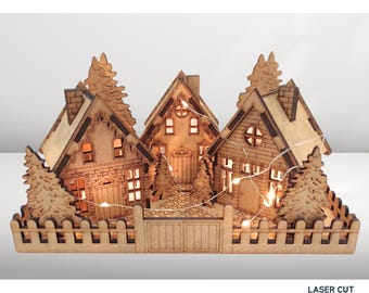 May include: A laser-cut wooden village scene featuring three houses, trees, and a fence. The houses have lit windows and are connected by a string of lights. The scene is made of light brown wood.
