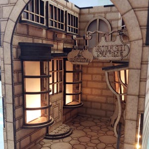 May include: A miniature wooden model of a shop with a sign that reads "Witch's Store" and another sign that reads "Hatmaker". The shop has a brick facade and a cobblestone floor. The windows are lit with warm light.