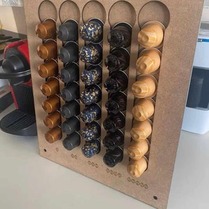 May include: A wooden wall-mounted organiser with rows of coffee pods. The pods are in various colours, including gold, black, and blue. The organiser has a simple design and is a great way to store and display coffee pods.