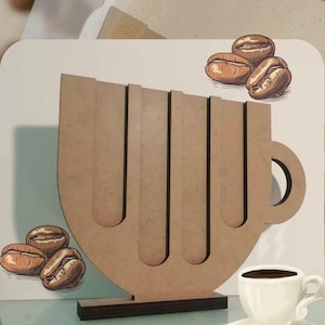 May include: A wooden coffee cup holder with four slots for coffee pods. The holder is shaped like a coffee cup with a handle. There are coffee beans drawn on the holder and a small cup of coffee on the table.
