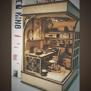 May include: A miniature wooden diorama of a writer's study, featuring a desk with a typewriter, bookshelves, and decorative elements. The diorama is set inside a small room with a detailed interior, including a rocking chair and artwork.