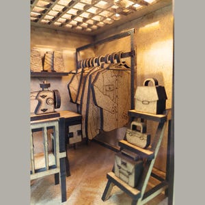 May include: A miniature wooden model of a tailor shop with a sewing machine, fabric rolls, shirts hanging on a rack, and purses on a shelf.