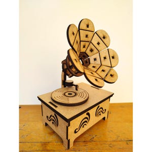 May include: A wooden model of a vintage gramophone. The model features a detailed speaker horn, a turntable, and a decorative base with curved accents. The gramophone is made of light brown wood with dark brown edges.