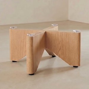 May include: A light-coloured wooden table base with a cross-shaped design. The base has four legs, each with a clear, circular foot and a black, circular base. The wood grain is visible, and the base is designed to support a tabletop.