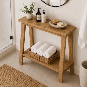 May include: A light brown wooden console table with an A-frame design, featuring a top shelf holding a small potted plant, two pump bottles, and a stone dish with a brush and soap. A woven basket on the lower shelf contains three white towels.
