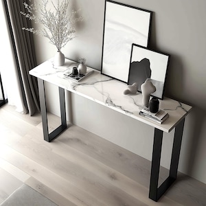 May include: A white marble console table with black metal legs. The table is decorated with black and white artwork, a vase with flowers, and decorative objects.