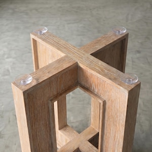 May include: Close-up of a light-colored wooden table base with an X-shaped design. The base has a natural wood grain finish and clear, circular suction cups on each corner. The table base is sturdy and modern.