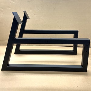 May include: Two black metal table legs with a rectangular frame and angled supports. The legs are made of sturdy metal and have a black powder coat finish.