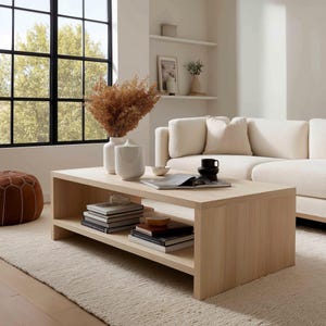 May include: A light oak coffee table with a lower shelf, displaying books and ornaments. Two white vases with dried flowers are on top, alongside a black mug and a bowl. The table is in a living room with a white sofa and a rug.