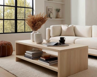 May include: A light oak coffee table with a lower shelf, displaying books and ornaments. Two white vases with dried flowers are on top, alongside a black mug and a bowl. The table is in a living room with a white sofa and a rug.