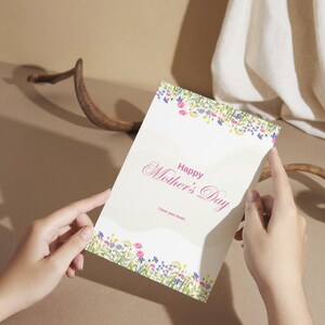 Op de afbeelding: Een Moederdagkaart die door handen wordt vastgehouden. De kaart toont de tekst "Happy Mother's Day" in een scriptlettertype, met "I love you mom" eronder. Bloemenontwerpen in roze, blauw en geel sieren de boven- en onderranden van de kaart.