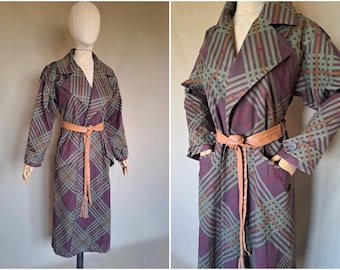 Op de afbeelding: Een vintage trenchcoat in een paars en groen ruitpatroon, tentoongesteld op een mannequin. De jas heeft een bruine riem, lange mouwen en een klassieke kraag. De jas is knielang en heeft zakken.