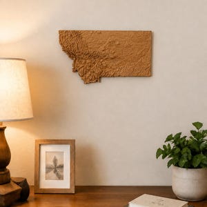 May include: A rectangular wall-mounted relief map of the state of Montana, rendered in a solid tan color. The map features a detailed, three-dimensional topographical surface showing the varied terrain, mountain ranges, and valleys of the region.