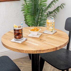 May include: A round wooden table with a black metal base, two black chairs, and two drinks on white coasters. A small white bowl with snacks sits on a white coaster in the centre of the table.