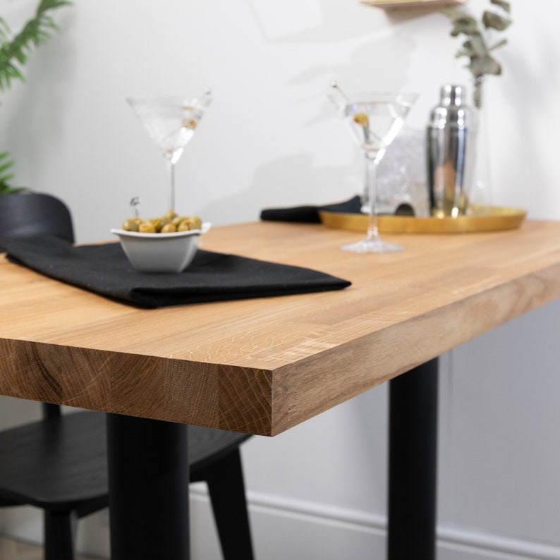 Pu&ograve; includere: Primo piano di un piano da tavolo in legno chiaro con gambe nere. Il tavolo &egrave; apparecchiato con un tovagliolo nero, una piccola ciotola di olive verdi e due bicchieri da martini.