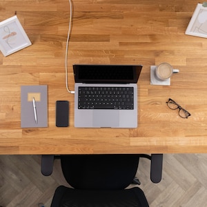 Pu&ograve; includere: Vista dall'alto di una scrivania in legno con un laptop, un quaderno, una penna, un telefono, una tazza di caff&egrave; e un paio di occhiali. La scrivania &egrave; anche dotata di una sedia.